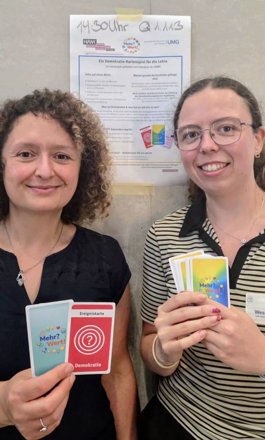 Die Gründerinnen Diana Heiman (links) und Pia Wessendorf (rechts) von Mehr?Wert! präsentieren lächelnd die Spielkarten vor einem Plakat zum Demokratie-Lernspiel.